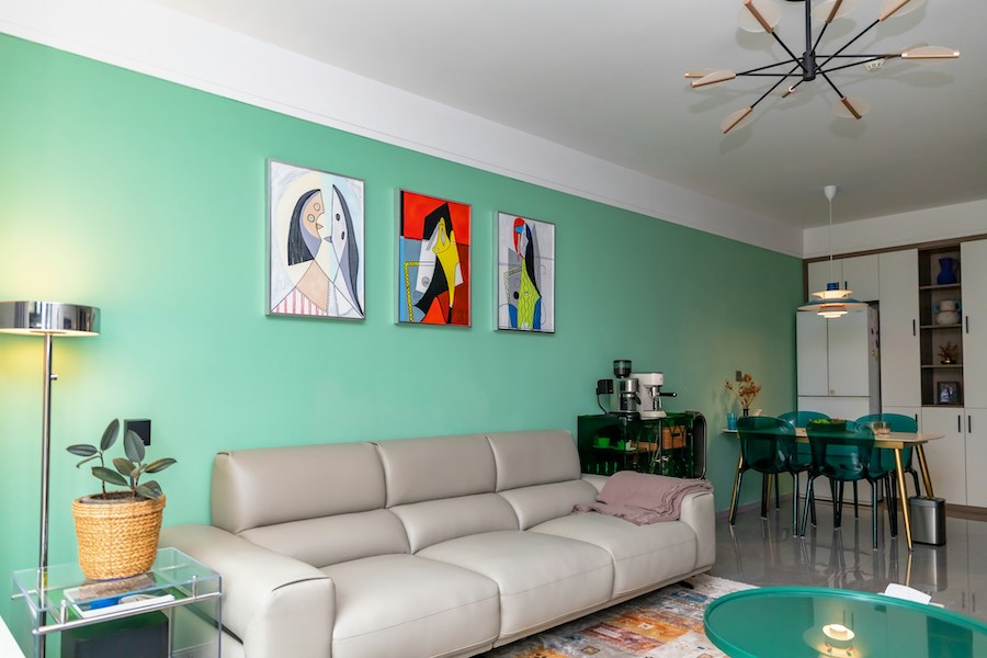 Интерьер кухни-гостиной в изумрудных тонах, обеденный стол, пластиковые стулья, столик с кофемашиной, диван, журнальный стол с подсветкой, постеры на стене, торшер, растение в плетенном кашпо