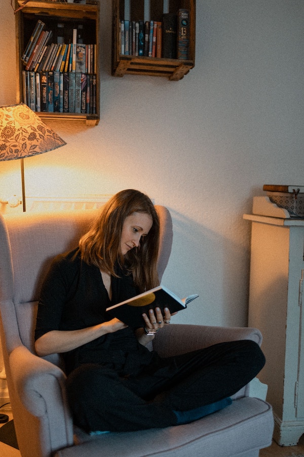 Женщина читает книгу под лампой, сидя на диване