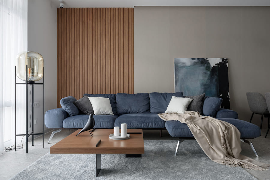 Разнообразие текстур и цветовые акценты добавляют динамики и выразительности интерьеру Гостиная, деревянная панель на стене, синий диван с подушками и пледом, картина на полу, журнальный стол с декоративной птичкой, торшер на металлической стойке